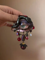 A textured iridescent hair brooch or barrette with a crackled purple-blue bead, red teardrop, glass beads, and a maroon bead with gold cap on a gold-toned chain, held by a hand with dark nail polish