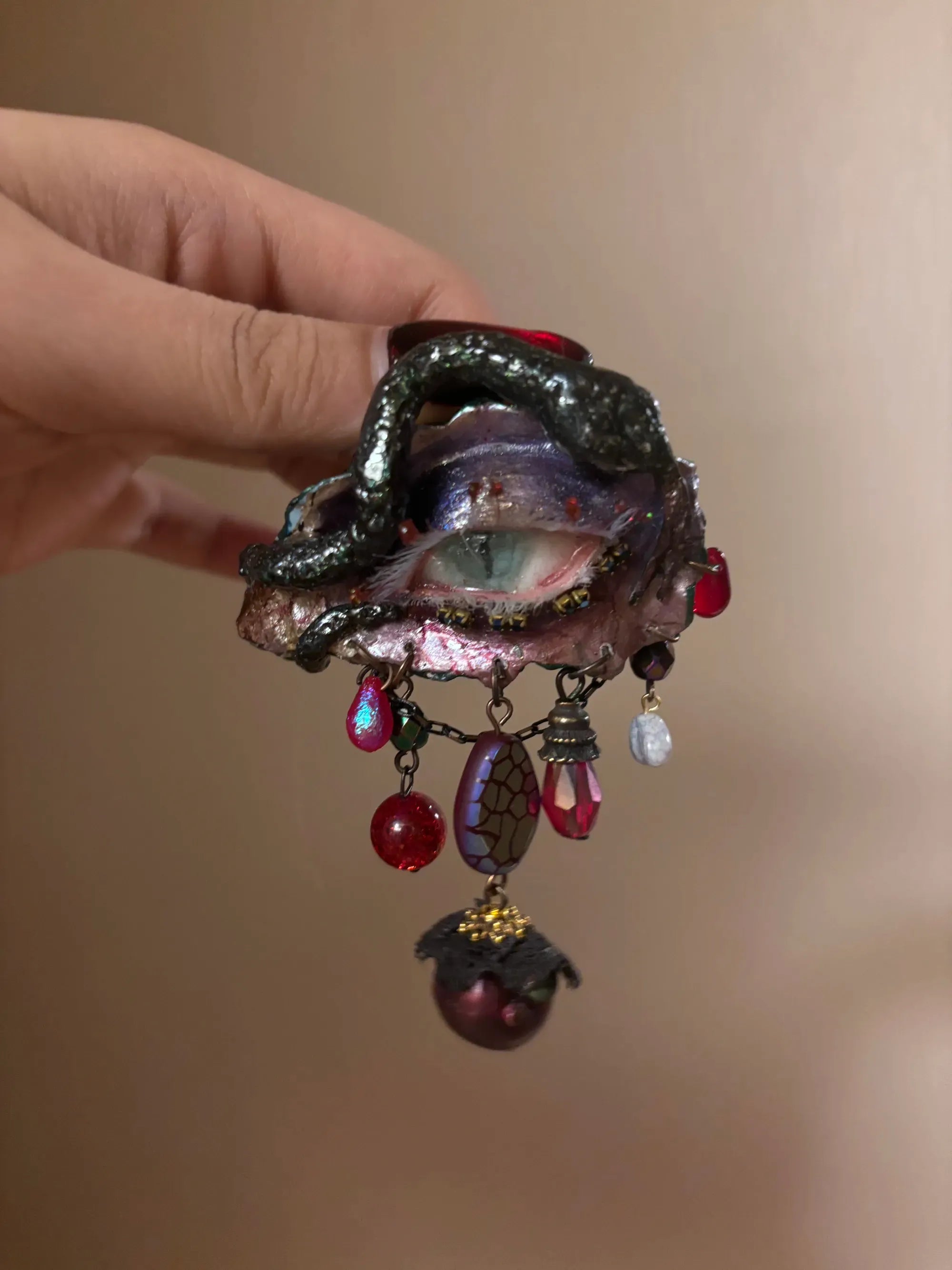 A textured iridescent hair brooch or barrette with a crackled purple-blue bead, red teardrop, glass beads, and a maroon bead with gold cap on a gold-toned chain, held by a hand with dark nail polish
