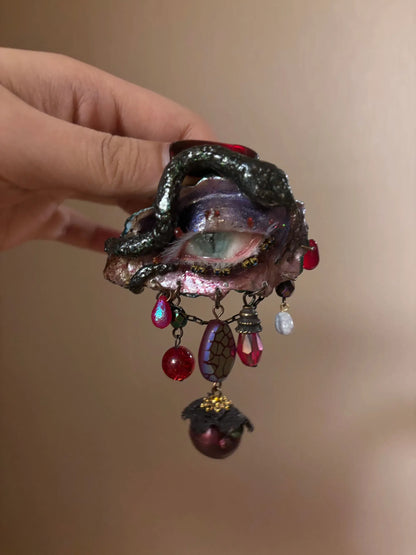A textured iridescent hair brooch or barrette with a crackled purple-blue bead, red teardrop, glass beads, and a maroon bead with gold cap on a gold-toned chain, held by a hand with dark nail polish