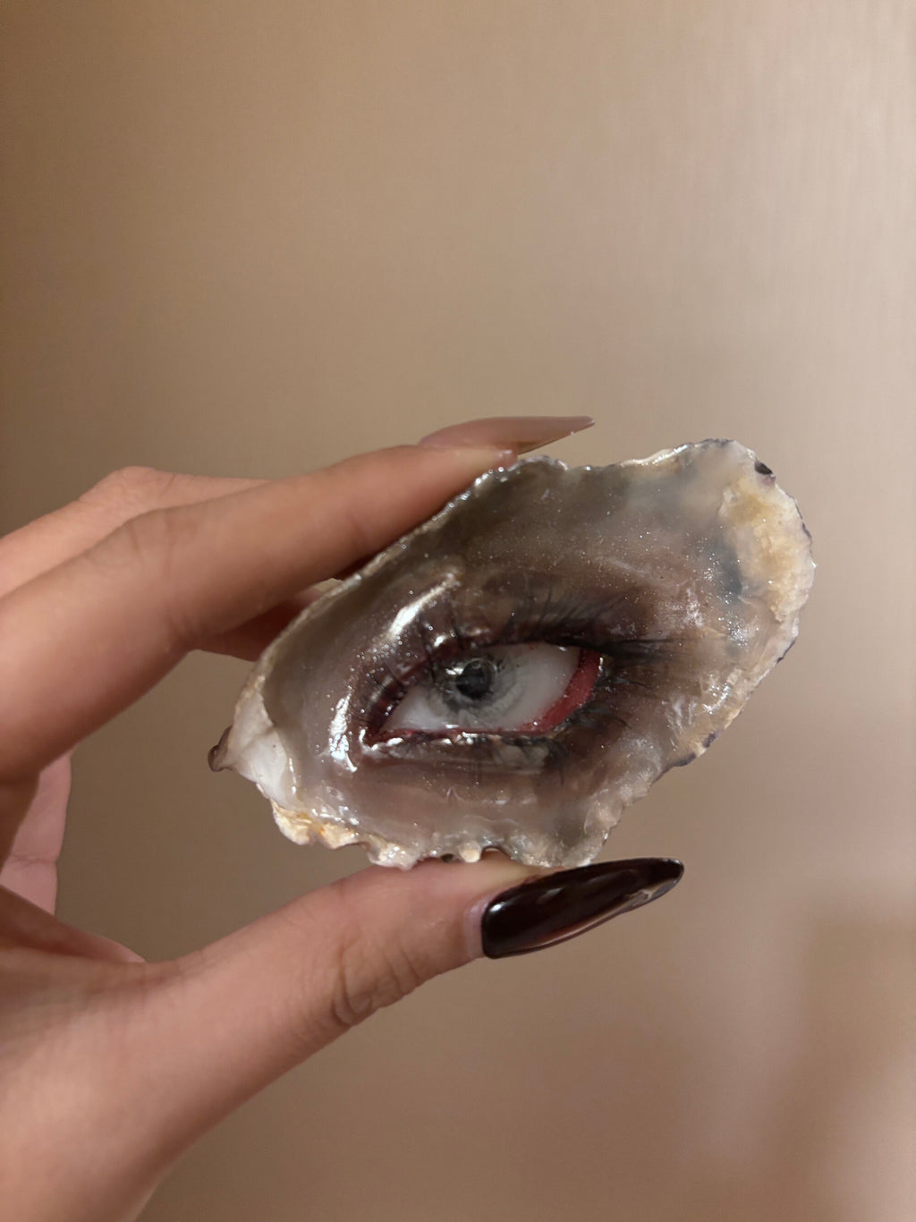 An irregular geode-like stone with a grey iris, black eyelashes, pink-red eyelids, resting on a reflective black glass surface with a mirror reflection beneath against a solid black background