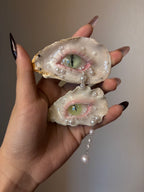 A decorated oyster shell with a cat-eye design, green iris, vertical slit pupil, rhinestone-adorned silver eyelid, white pearl tears, and dangling pearl chain, held by a hand with glossy black nail art against a grey background