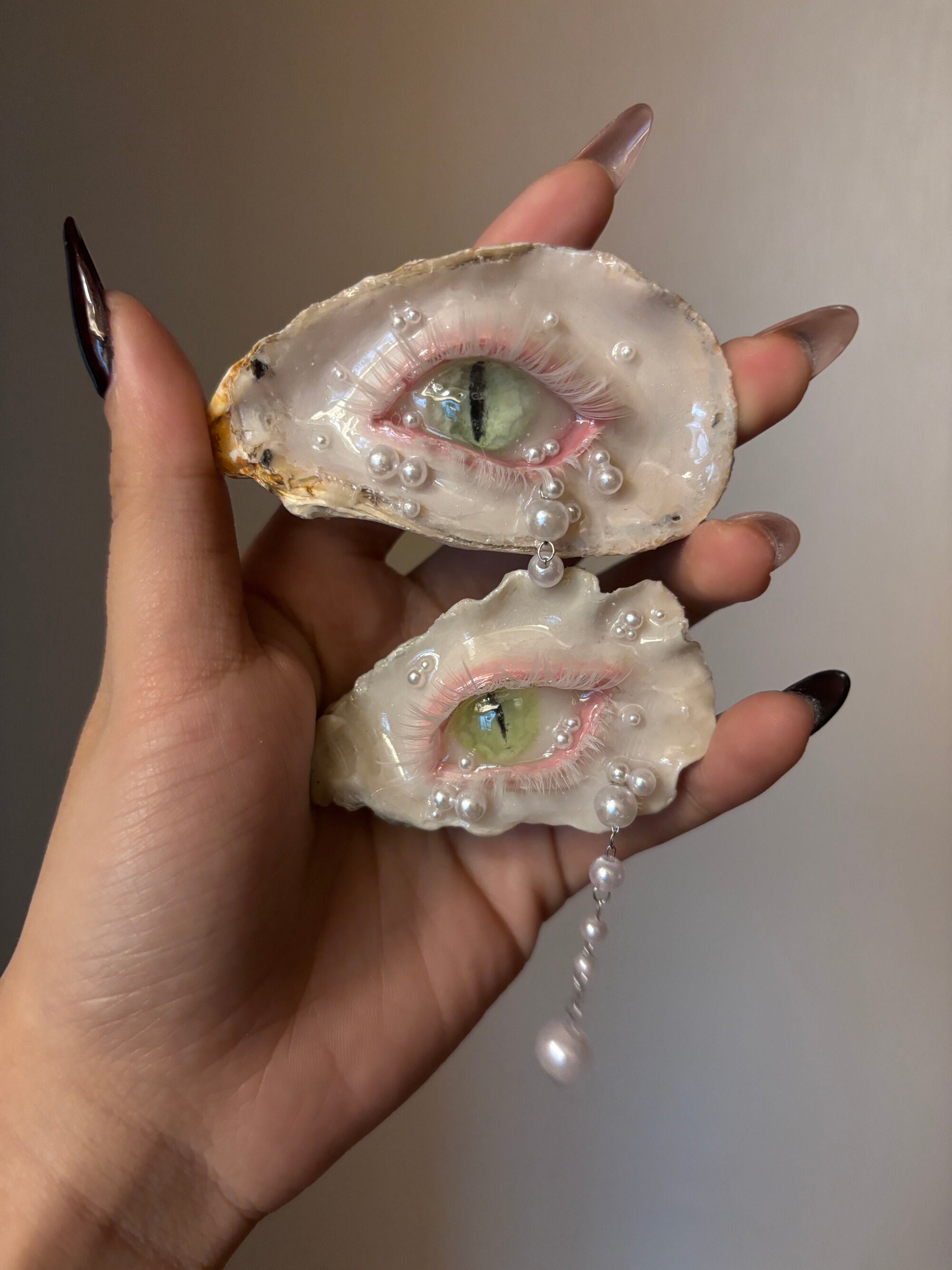 A decorated oyster shell with a cat-eye design, green iris, vertical slit pupil, rhinestone-adorned silver eyelid, white pearl tears, and dangling pearl chain, held by a hand with glossy black nail art against a grey background