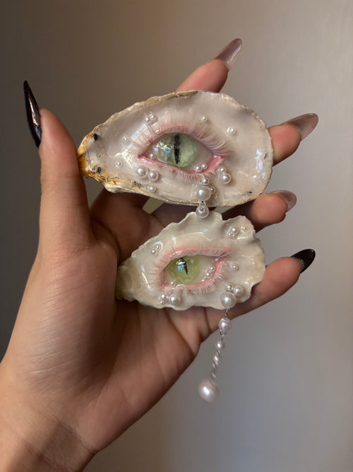 A decorated oyster shell with a cat-eye design, green iris, vertical slit pupil, rhinestone-adorned silver eyelid, white pearl tears, and dangling pearl chain, held by a hand with glossy black nail art against a grey background