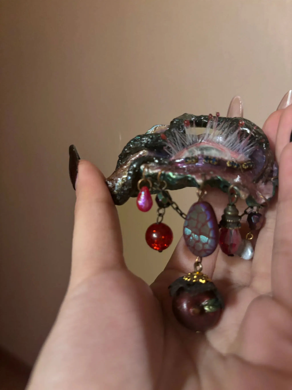 A textured metallic green patina brooch with dangling red teardrop beads, faceted purple bead, maroon bead with black lace top, and red glitter bead on an antique gold chain against a beige background