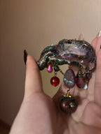 A textured metallic green patina brooch with dangling red teardrop beads, faceted purple bead, maroon bead with black lace top, and red glitter bead on an antique gold chain against a beige background