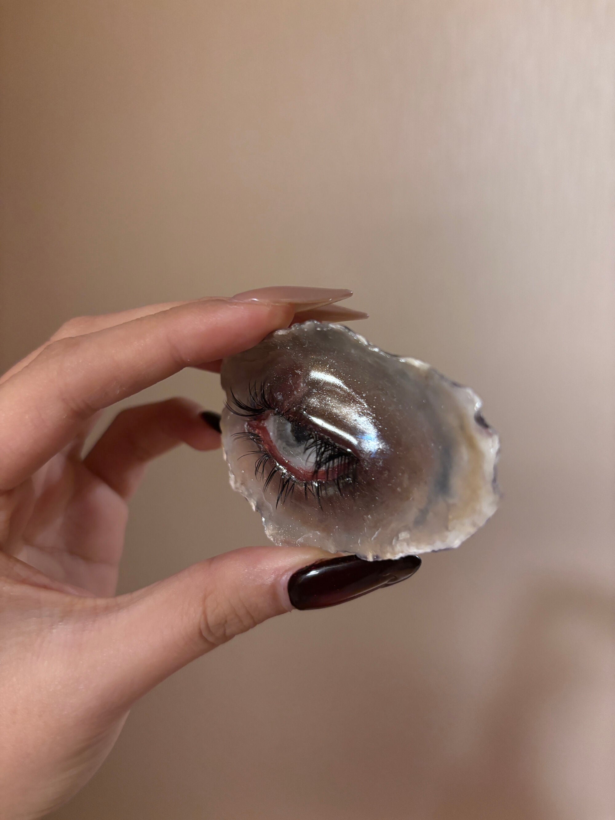 A hand holding a resin or natural stone piece with a blue-grey iris, black eyelashes, red-toned eyelid, and glossy surface against a beige background, displaying a surreal geode-eye aesthetic