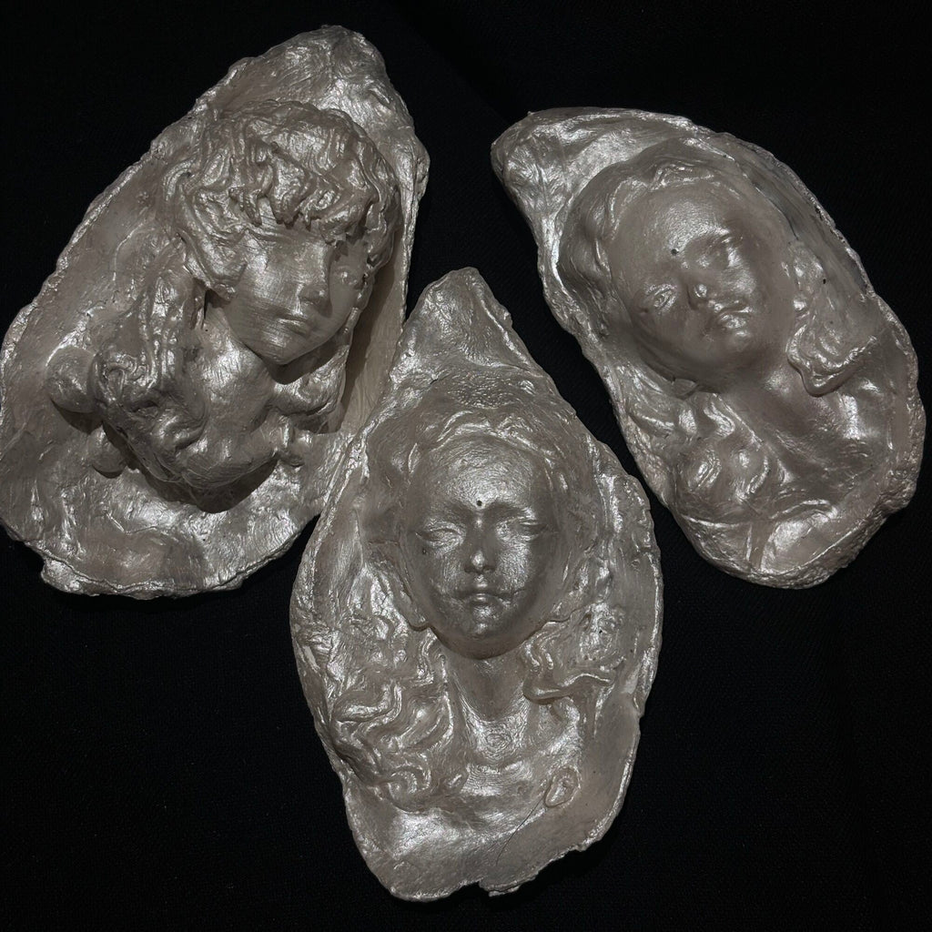 A sculptural silver-toned baroque pearl pendant depicting a serene woman's face with closed eyes and curled hair, suspended from a frayed ivory ribbon, shown against a dark background