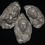 A sculptural silver-toned baroque pearl pendant depicting a serene woman's face with closed eyes and curled hair, suspended from a frayed ivory ribbon, shown against a dark background