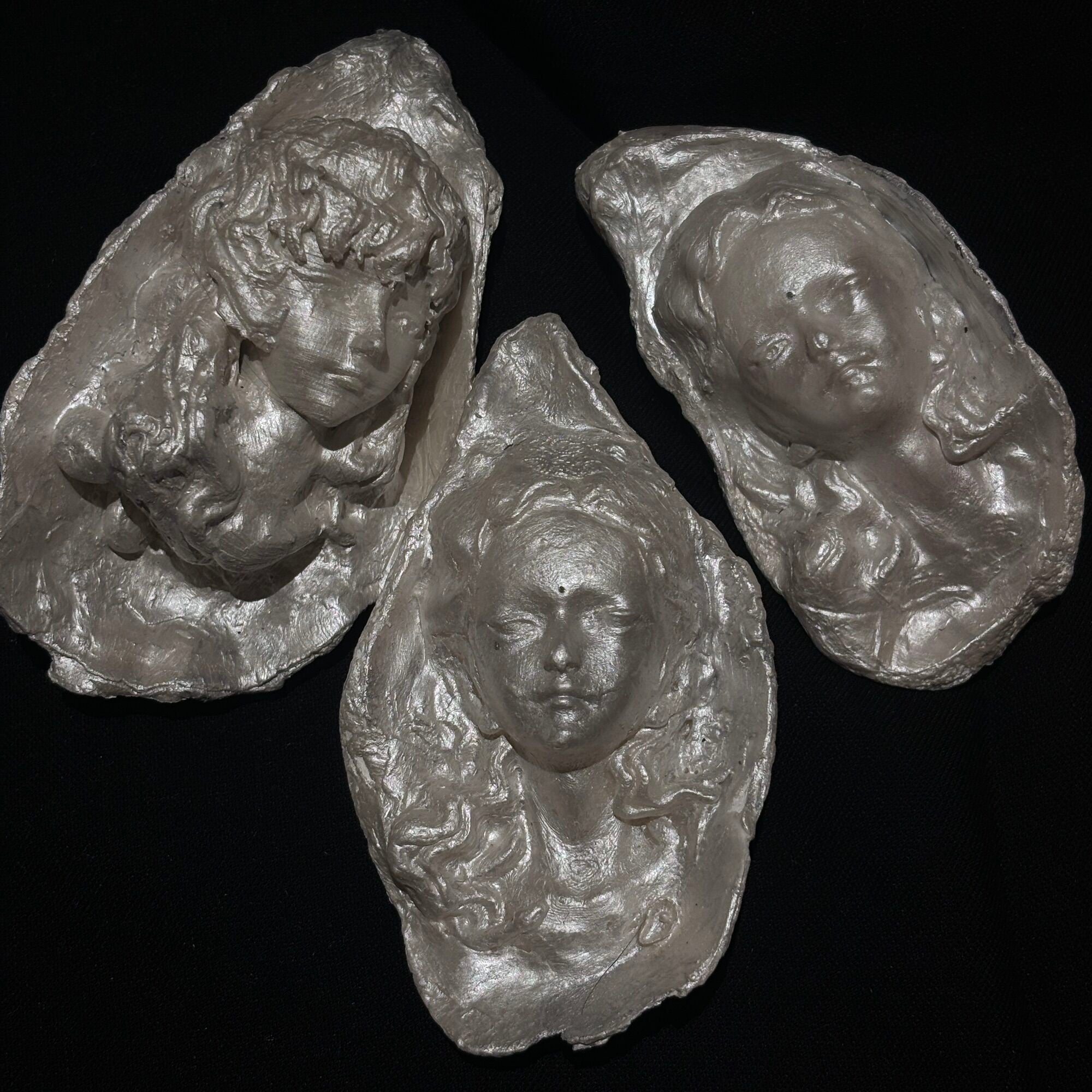 A sculptural silver-toned baroque pearl pendant depicting a serene woman's face with closed eyes and curled hair, suspended from a frayed ivory ribbon, shown against a dark background