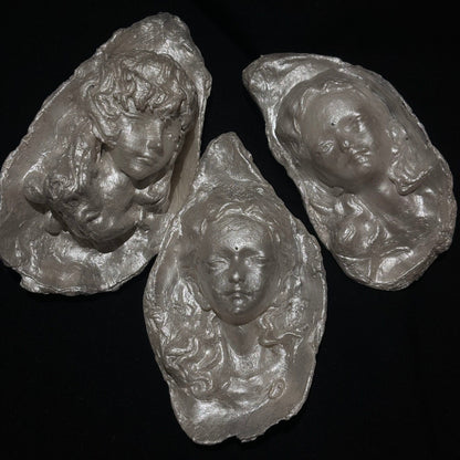 A sculptural silver-toned baroque pearl pendant depicting a serene woman's face with closed eyes and curled hair, suspended from a frayed ivory ribbon, shown against a dark background
