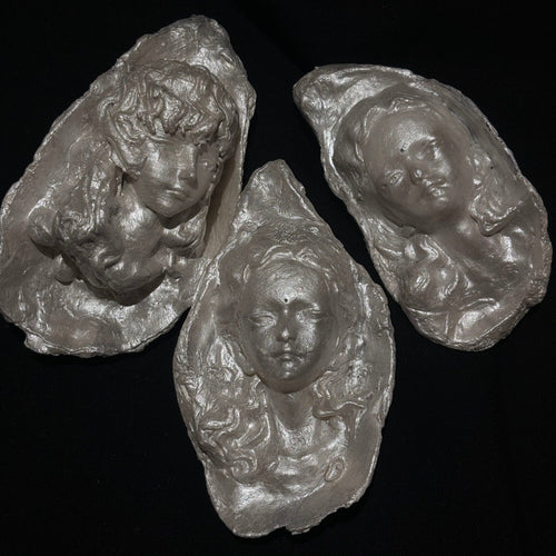 A sculptural silver-toned baroque pearl pendant depicting a serene woman's face with closed eyes and curled hair, suspended from a frayed ivory ribbon, shown against a dark background