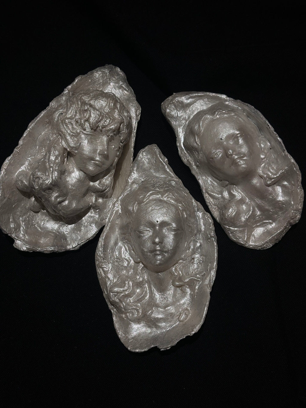A sculptural silver-toned pendant with a woman's face and embedded pearls resting on a hand against a dark background, emphasizing its wearable scale and artisan texture