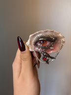 An oyster shell with a green cat-eye iris, vertical slit pupil, pink eyelids, white pearl tears, and a dangling pearl chain, held in a hand against a neutral grey background