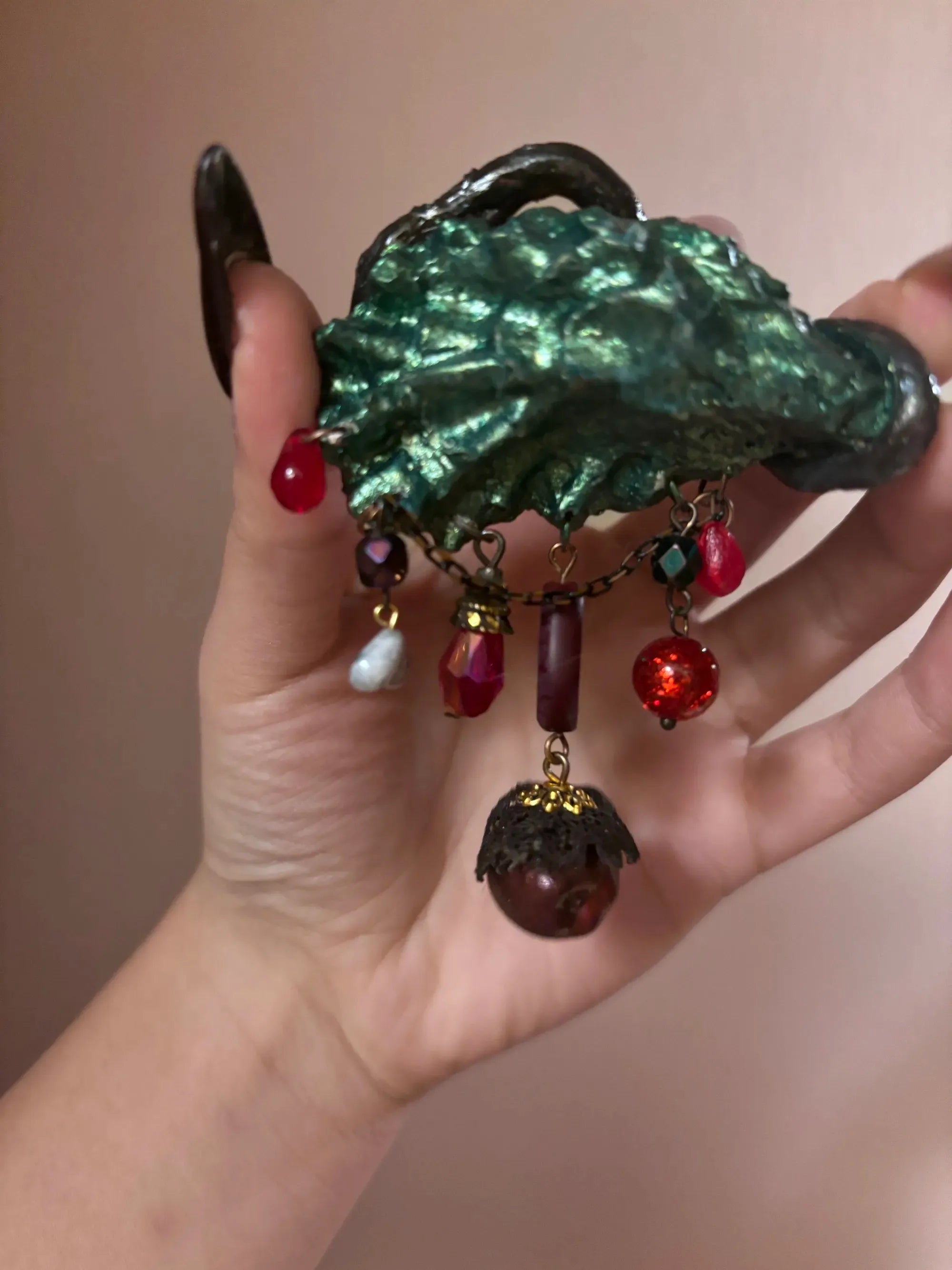 A gothic enamel serpent eye brooch with blue-green iris, pink eyelids, black vine surround, and cascading iridescent beads and charms against a solid black background