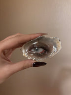 A glossy grey-brown shell or resin piece with a blue iris, red-toned eyelid, realistic black eyelashes, and a reflective surface held by a hand with dark maroon nail polish