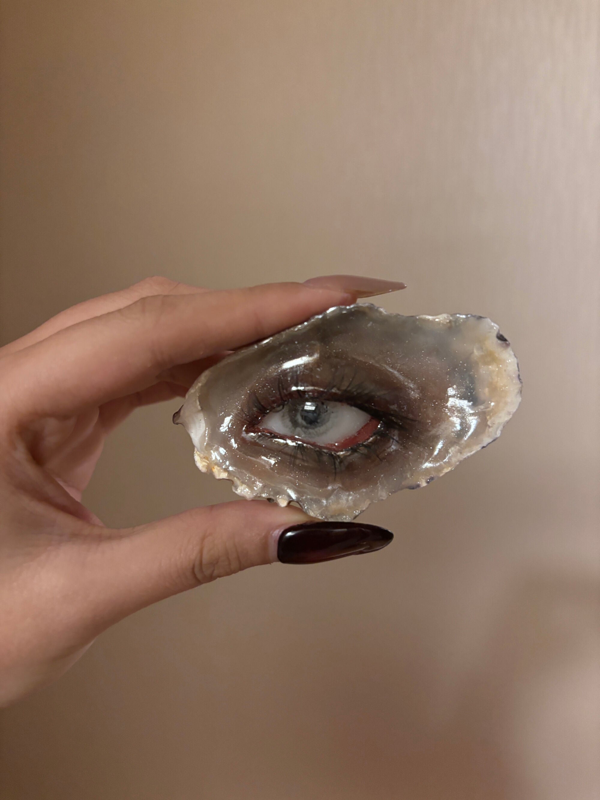 A glossy grey-brown shell or resin piece with a blue iris, red-toned eyelid, realistic black eyelashes, and a reflective surface held by a hand with dark maroon nail polish
