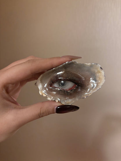 A glossy grey-brown shell or resin piece with a blue iris, red-toned eyelid, realistic black eyelashes, and a reflective surface held by a hand with dark maroon nail polish