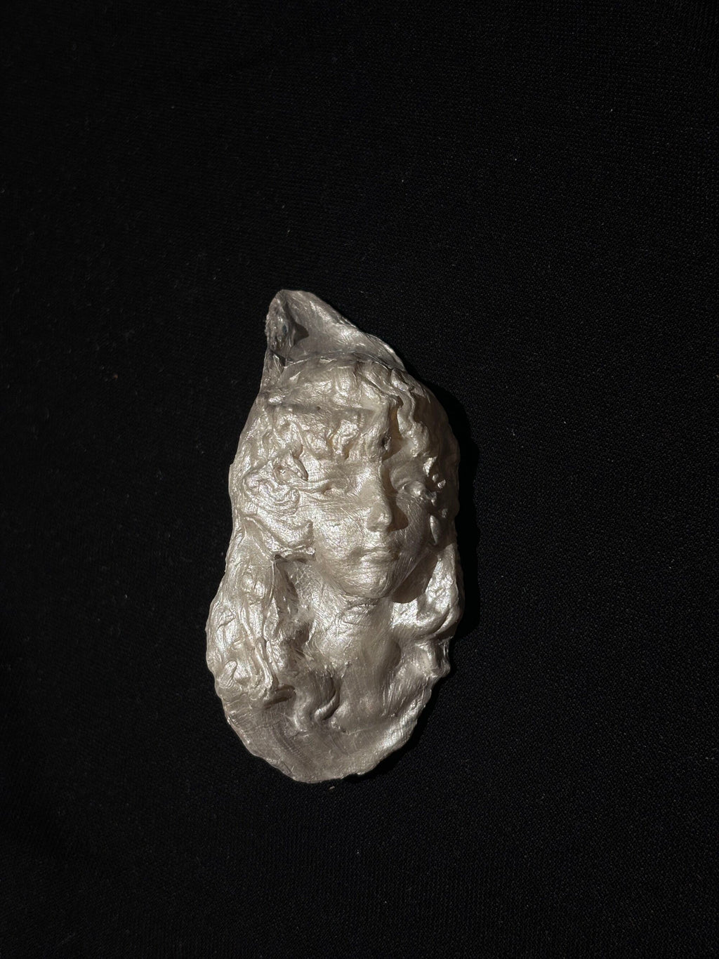 A sculptural silver-toned pendant with a woman's face and embedded pearls resting on a hand against a dark background, emphasizing its wearable scale and artisan texture