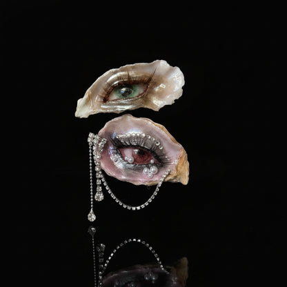 Two oyster shells transformed into surreal eye art pieces with green cat-eye irises, pink eyelids, white pearl tears, and a teardrop pearl chain, held by a hand against a beige striped wallpaper