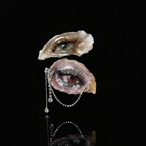 Two oyster shells transformed into surreal eye art pieces with green cat-eye irises, pink eyelids, white pearl tears, and a teardrop pearl chain, held by a hand against a beige striped wallpaper