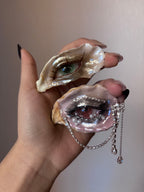 A hand wearing an oyster shell eye brooch with a green iris, brown eyelashes, and pearl accents, shown alongside a second shell with a green iris against a beige background