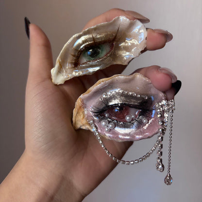 A hand wearing an oyster shell eye brooch with a green iris, brown eyelashes, and pearl accents, shown alongside a second shell with a green iris against a beige background