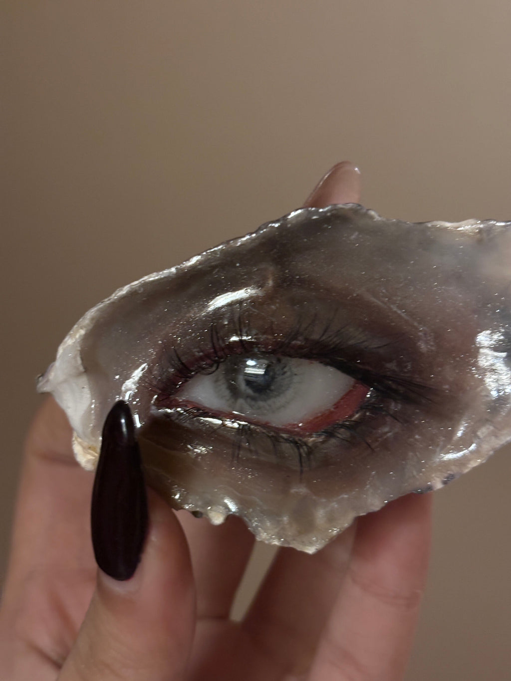 A hand with a dark burgundy nail holding a shell or resin eye piece featuring a blue-grey iris, red eyelid, and realistic eyelashes against a beige background