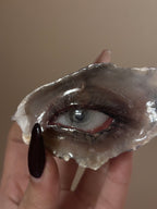 A hand with a dark burgundy nail holding a shell or resin eye piece featuring a blue-grey iris, red eyelid, and realistic eyelashes against a beige background