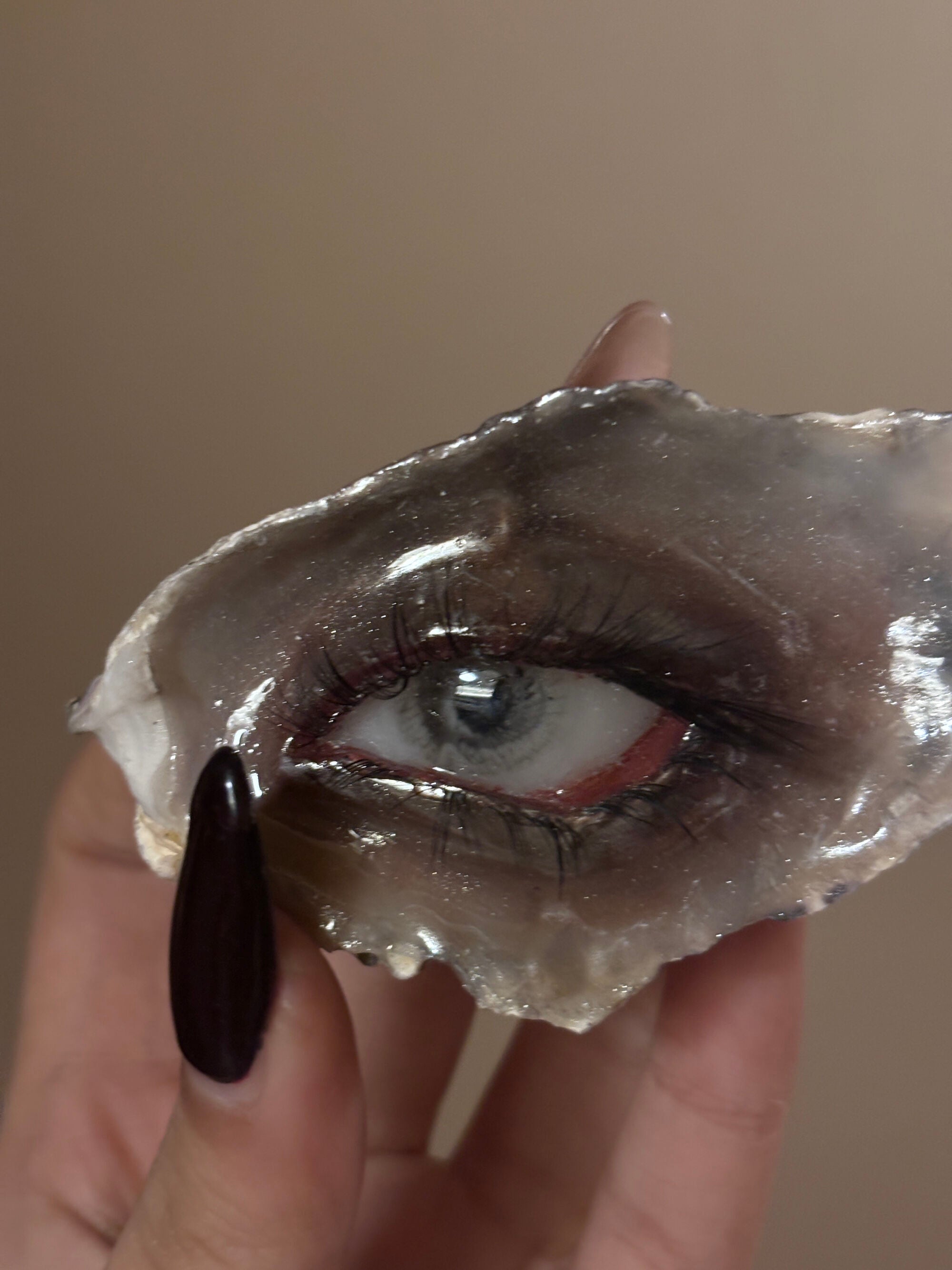 A hand with a dark burgundy nail holding a shell or resin eye piece featuring a blue-grey iris, red eyelid, and realistic eyelashes against a beige background