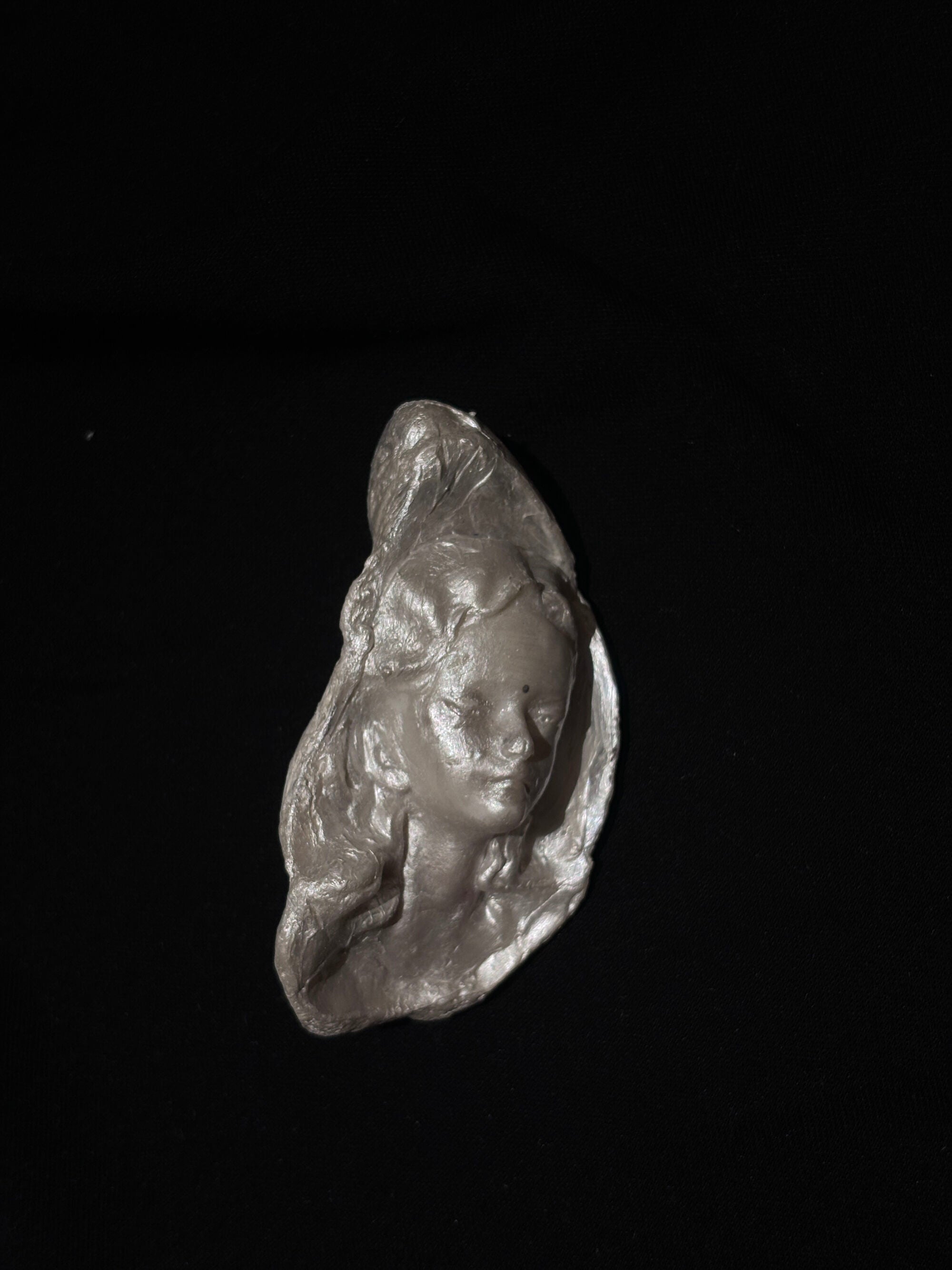 A baroque pearl pendant with a sculpted woman's face and organic irregular base, hung on a frayed ivory linen ribbon against a matte black background
