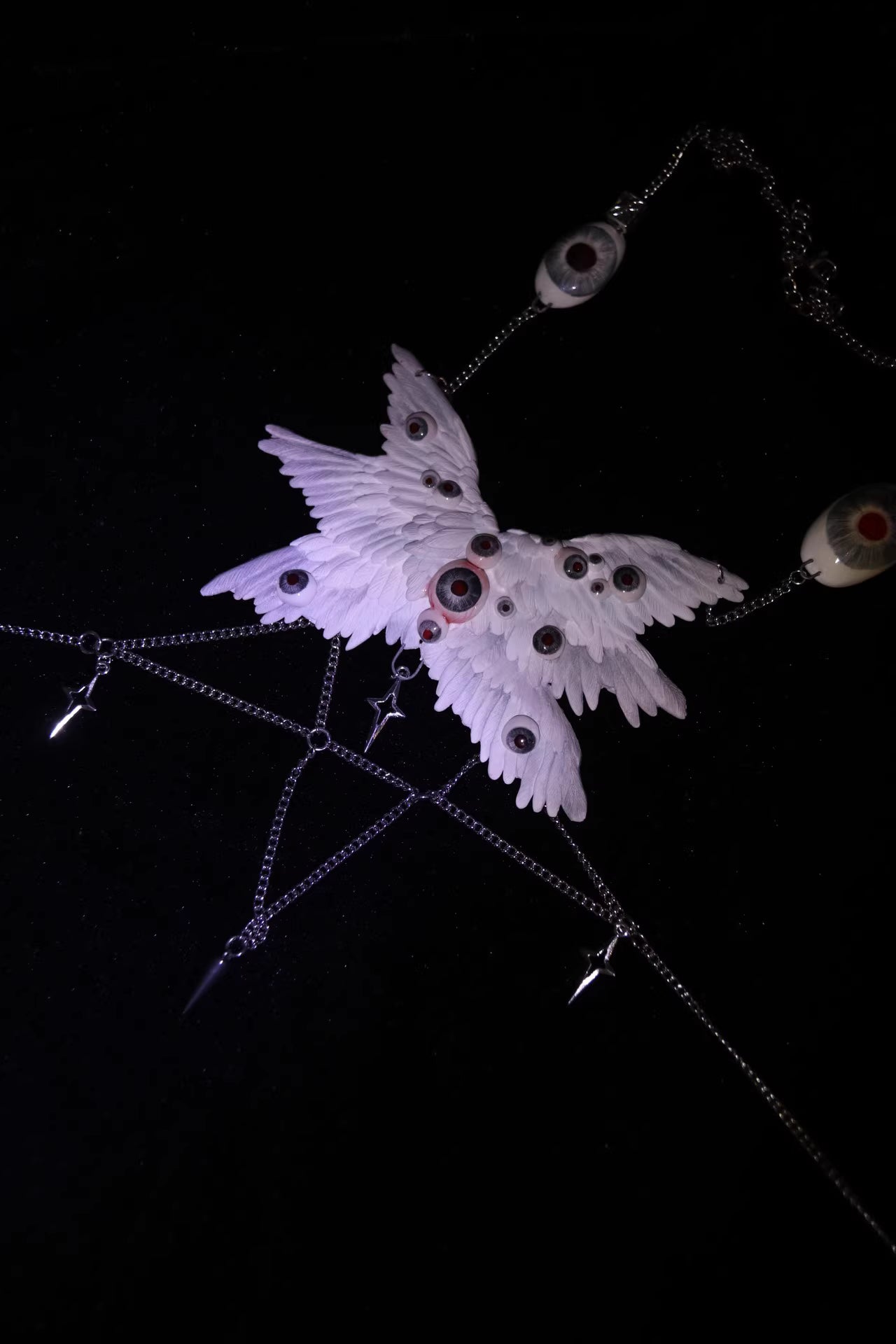 Macabre ceramic angel wing body chain relic against dark ground — white sculpted feather form emerges from shadow, glass eyeball nodes catching ambient light across the wing surface. The chain trails below like a severed tether.